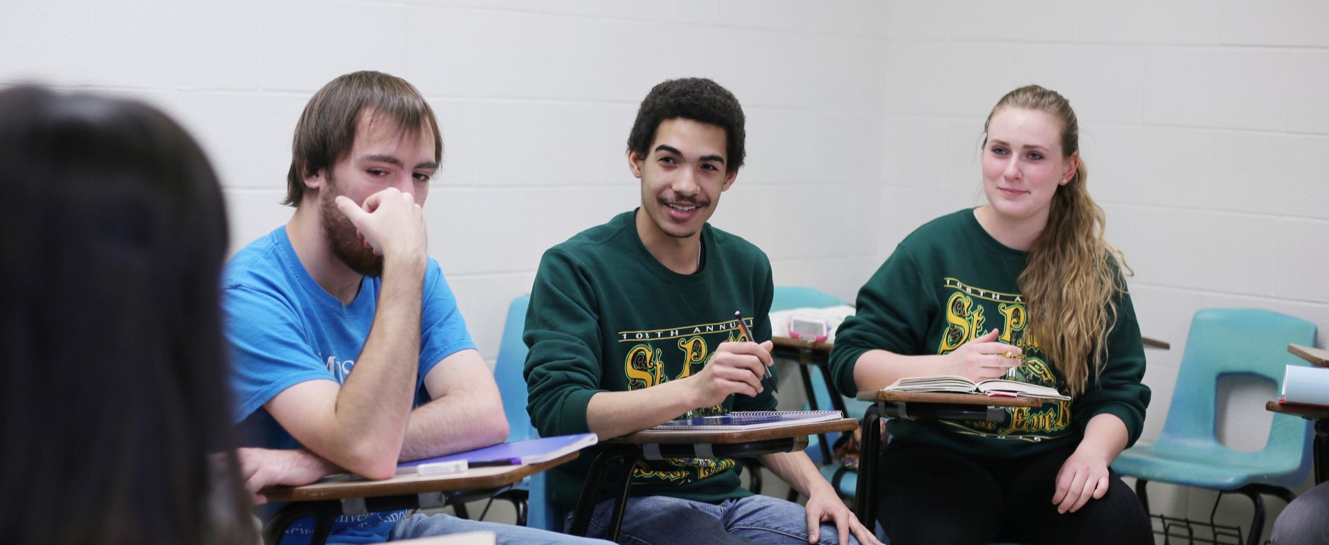 Three students sit in a row in classroom resks. The middle student is engaged and smiling with the instructor while the other two listen.